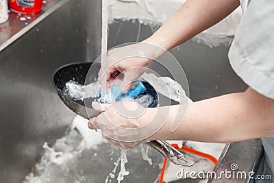 Closeup of dishwasher in uniform washes a pan with foam and a sponge under the tap water in a metal sink in the restaurantâ€™s Stock Photo